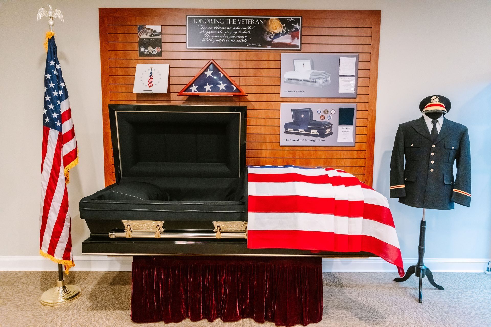 A funeral display featuring a black casket draped in an American flag, with a military uniform and a folded flag behind it.