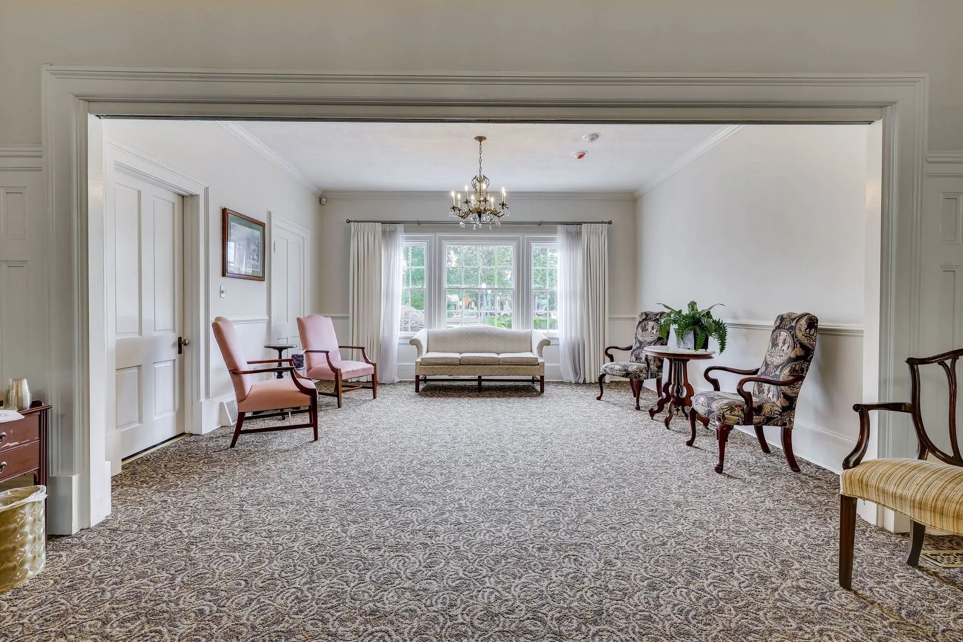 A spacious, bright room with patterned carpet features a white sofa, two pink chairs, and several patterned armchairs.