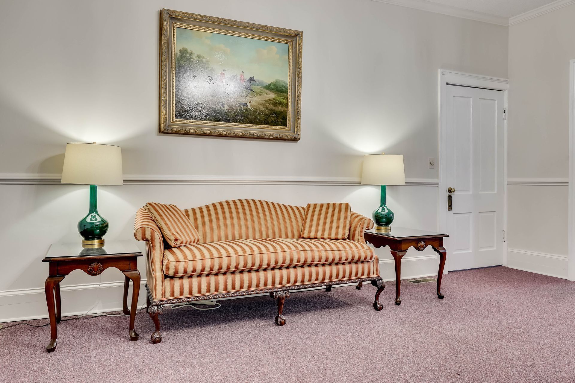 A striped couch sits between two side tables with lamps in front of a framed landscape painting on a neutral wall.