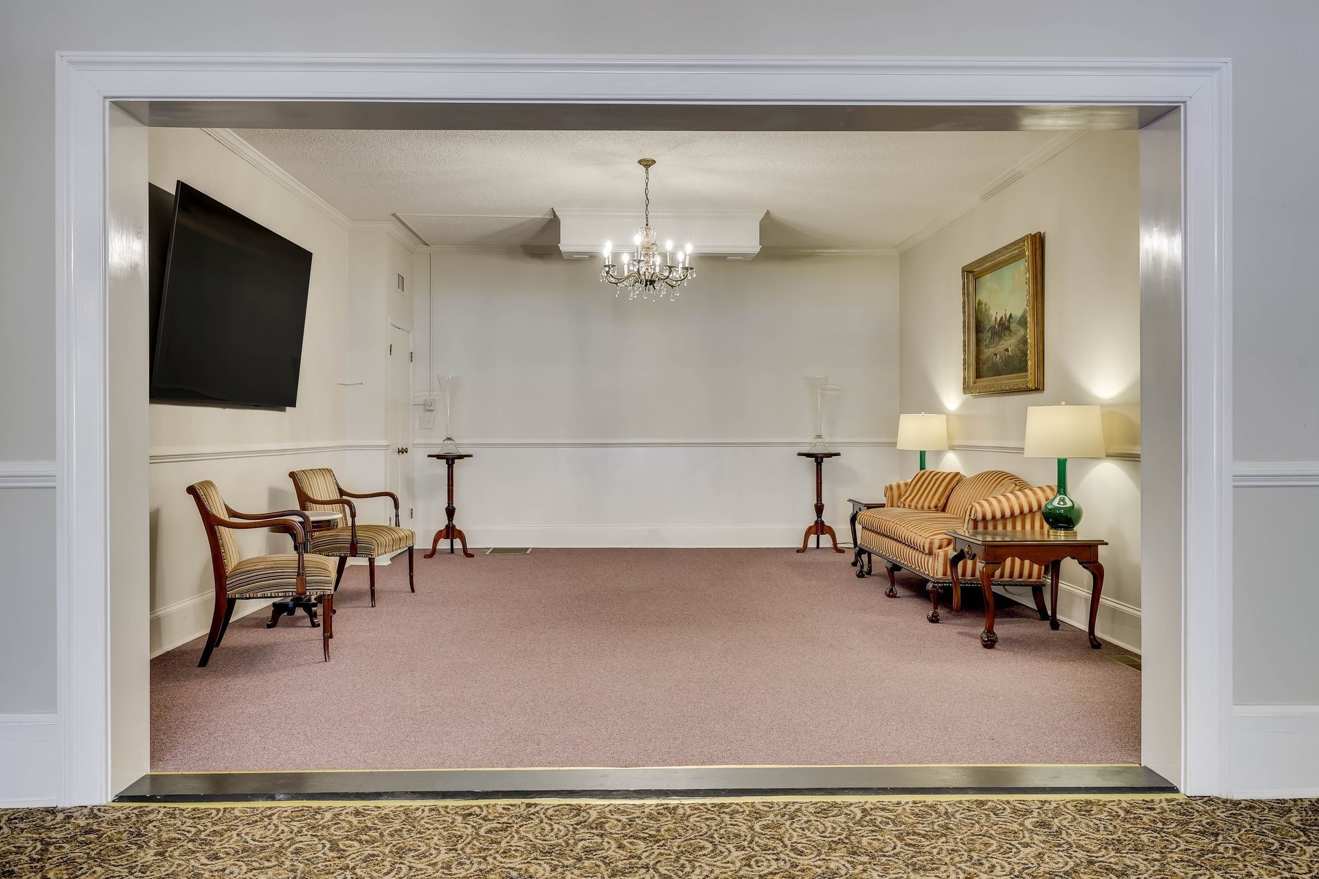 A formal room viewed through an archway, featuring two armchairs, a striped sofa, a chandelier, and a wall-mounted TV.