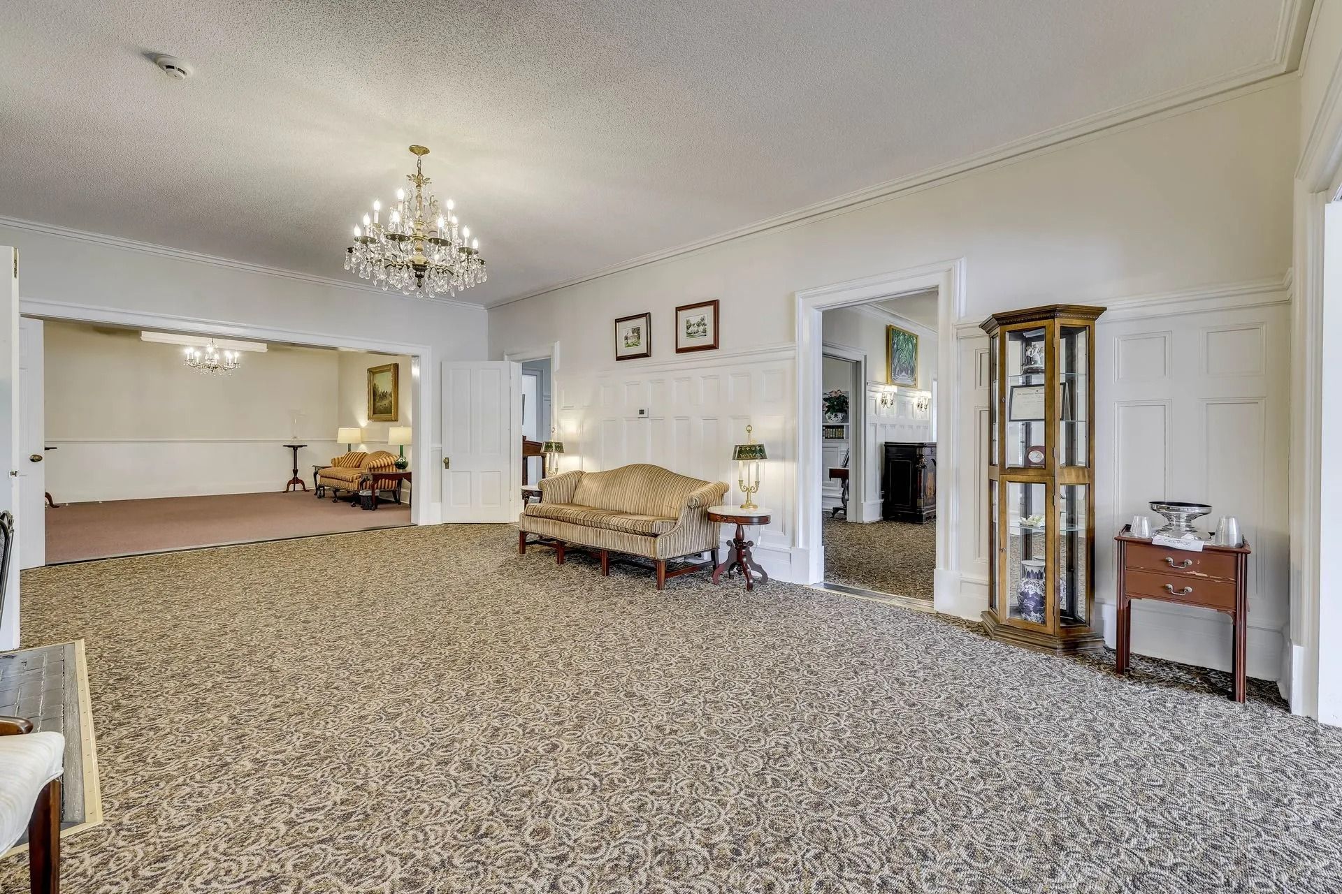 A brightly lit living room with patterned carpet, a beige sofa, a crystal chandelier, and a wood display cabinet.