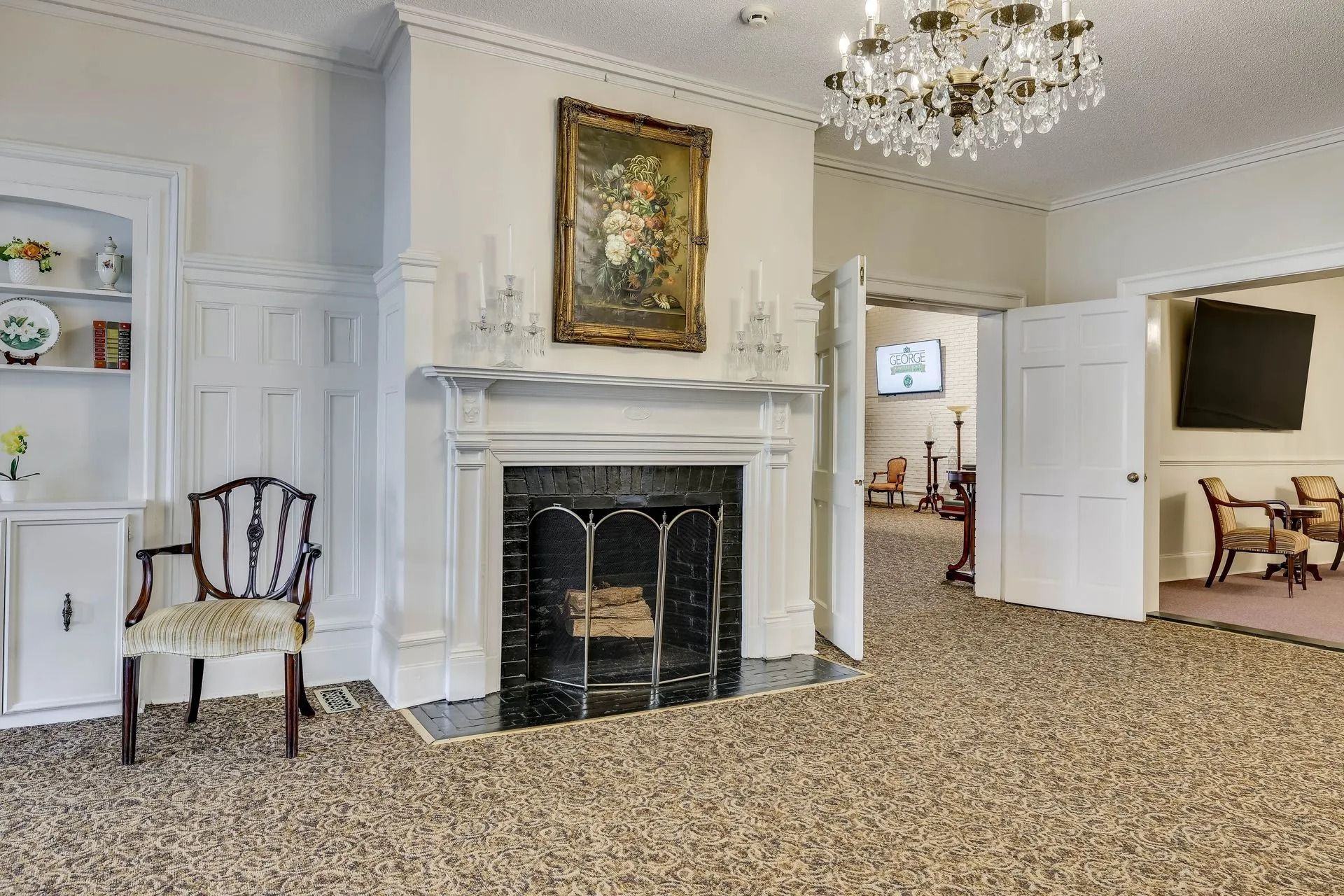 An elegant room with a central fireplace, an oil painting, a crystal chandelier, and a single chair on a patterned carpet.