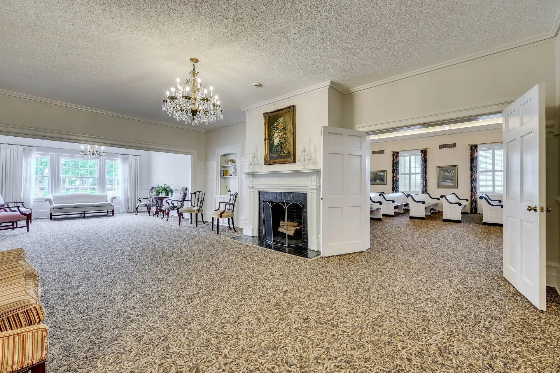 Large, carpeted room with a fireplace, ornate chandelier, and open doorways leading to an adjacent seating area.