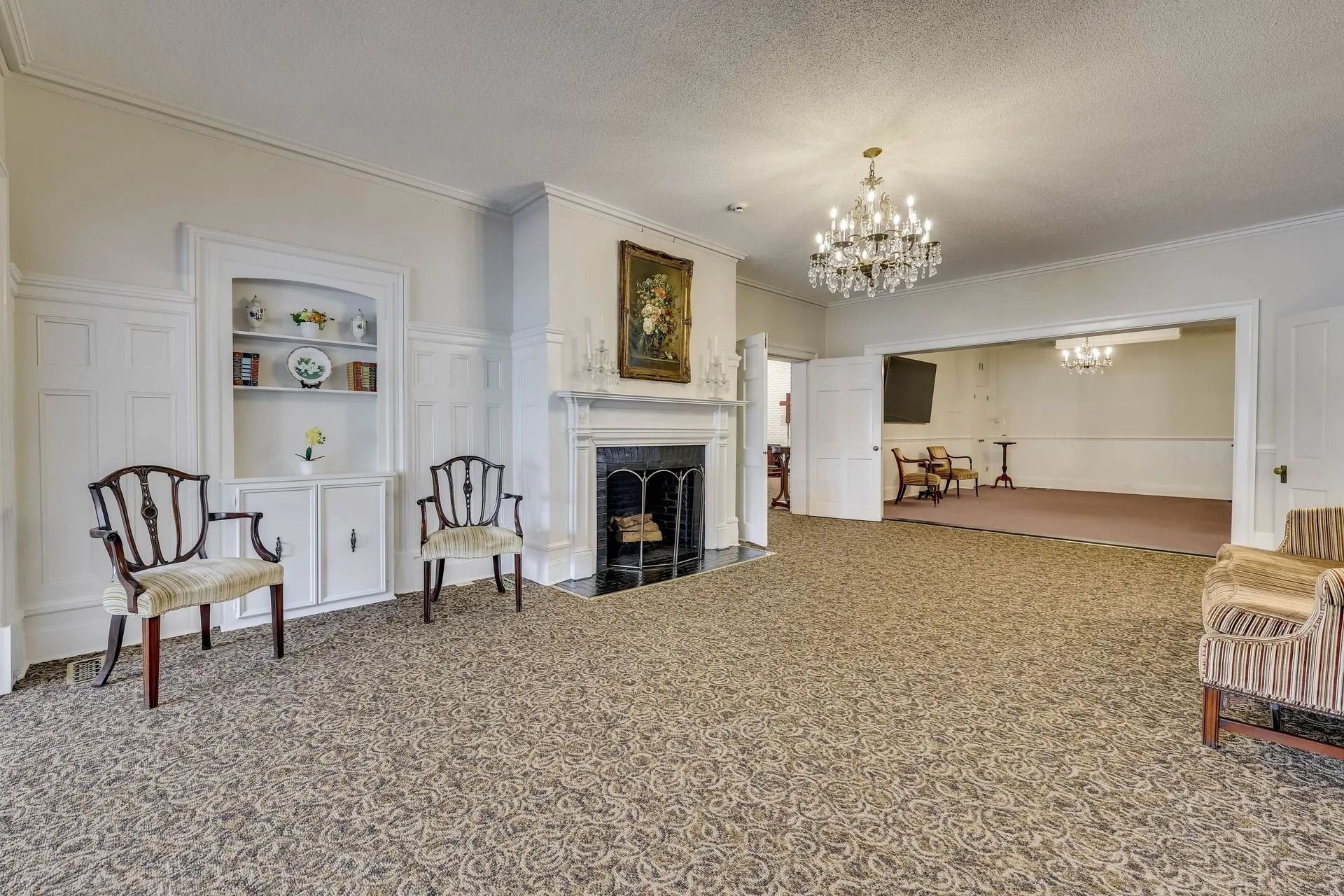 Living room with a fireplace, chandelier, built-in shelving, two chairs, and patterned carpeting leading to another room.