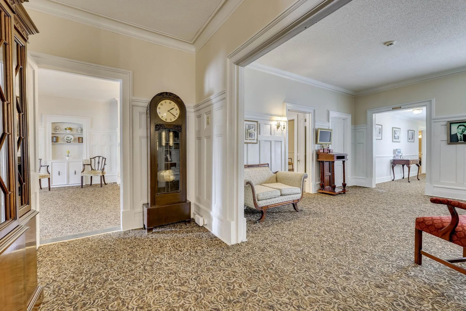 A spacious room with beige carpet, a grandfather clock, a light-colored settee, and doorways leading to other rooms.