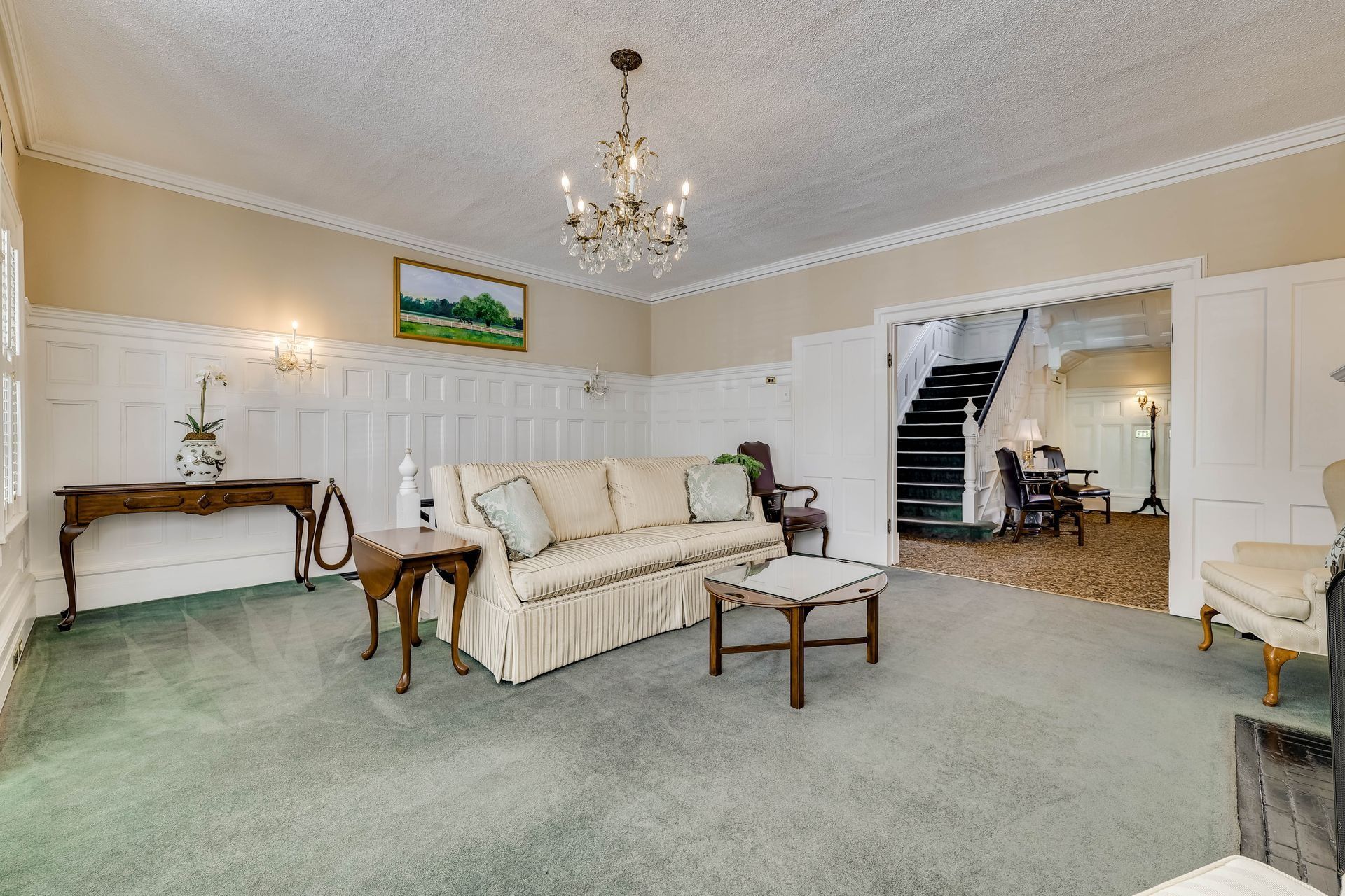 A living room with light walls, wainscoting, a beige sofa, green carpet, a chandelier, and a view into a hallway with stairs.