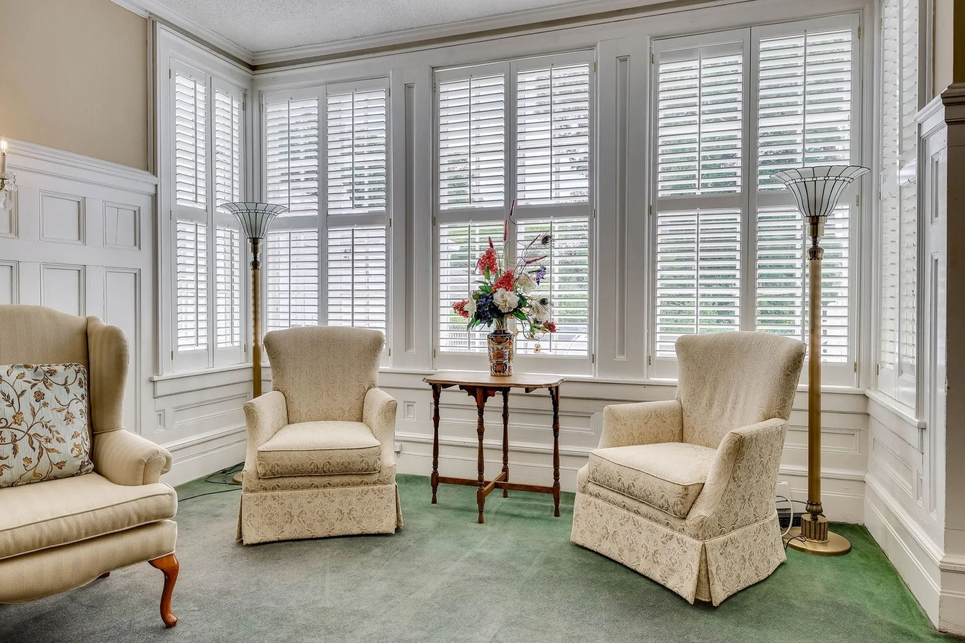 A sunroom featuring three beige wingback chairs, a central wooden pedestal table with a floral arrangement, and lamps.