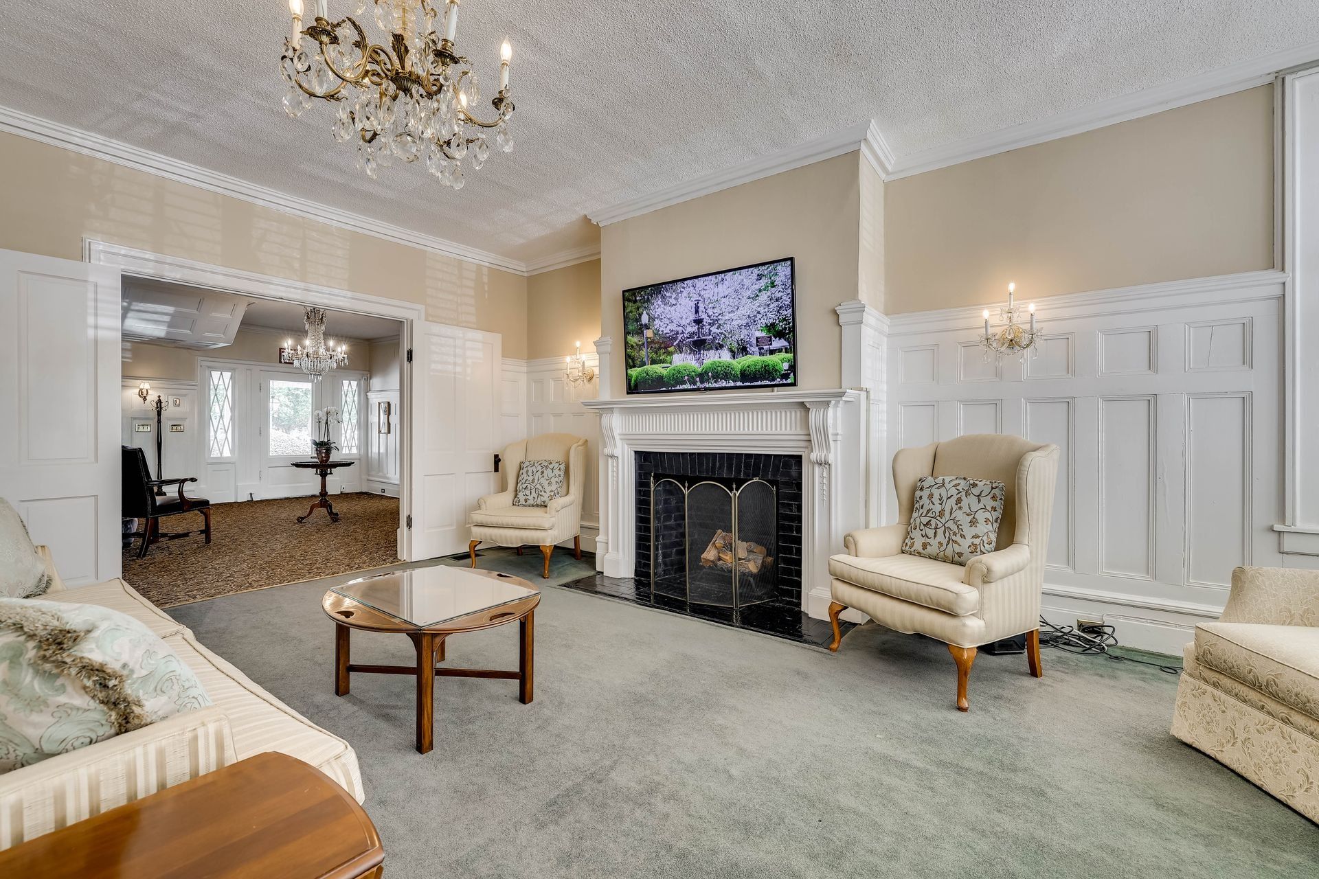 A living room featuring a fireplace with a mounted television, two cream armchairs, a coffee table, and chandeliers.