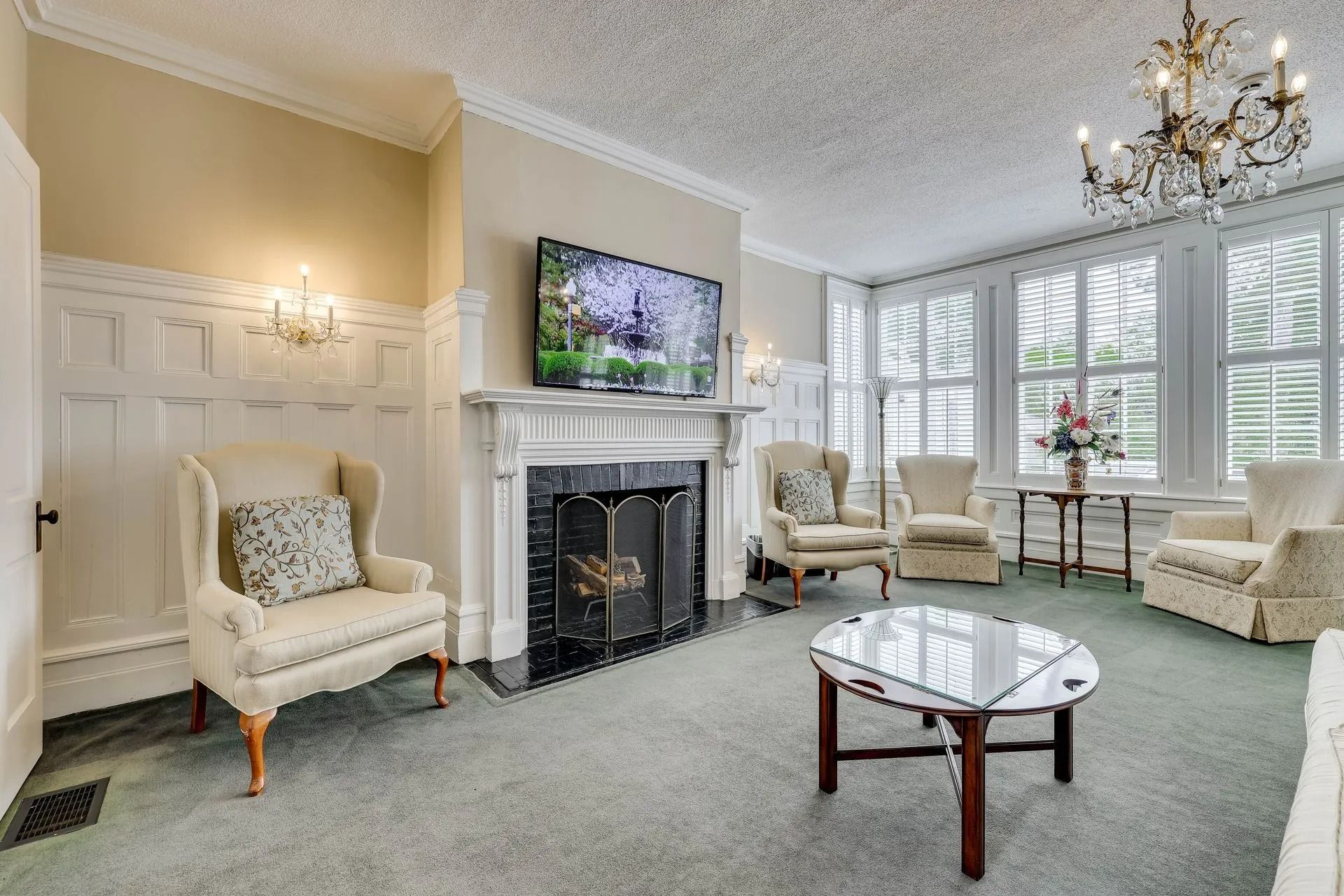 A living room featuring white wainscoting, a fireplace with a mounted television, a glass coffee table, and armchairs.