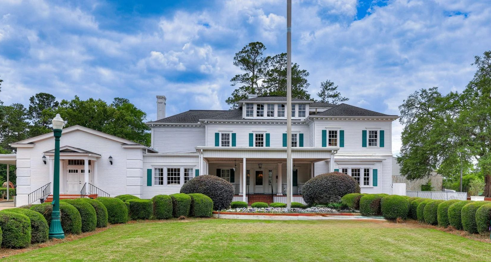 A white two-story historic house with green shutters, a front porch, and a manicured lawn under a partly cloudy blue sky.