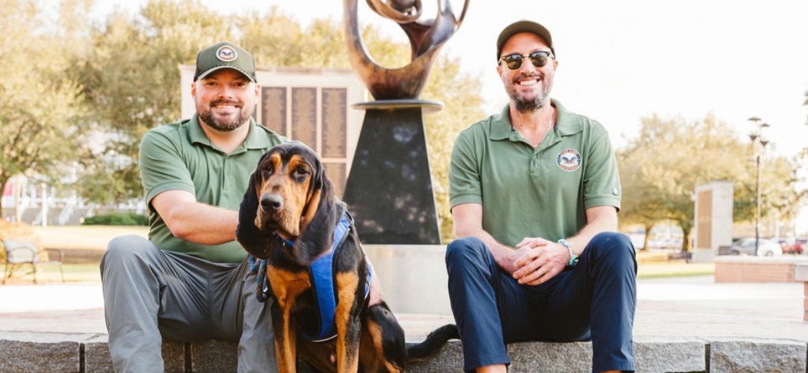 Two men sit with a black-and-tan dog on a bench outdoors, with a sculpture behind them.