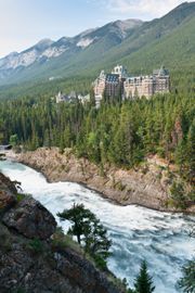 Castle on a forested riverbank with mountains in the background and whitewater rushing below