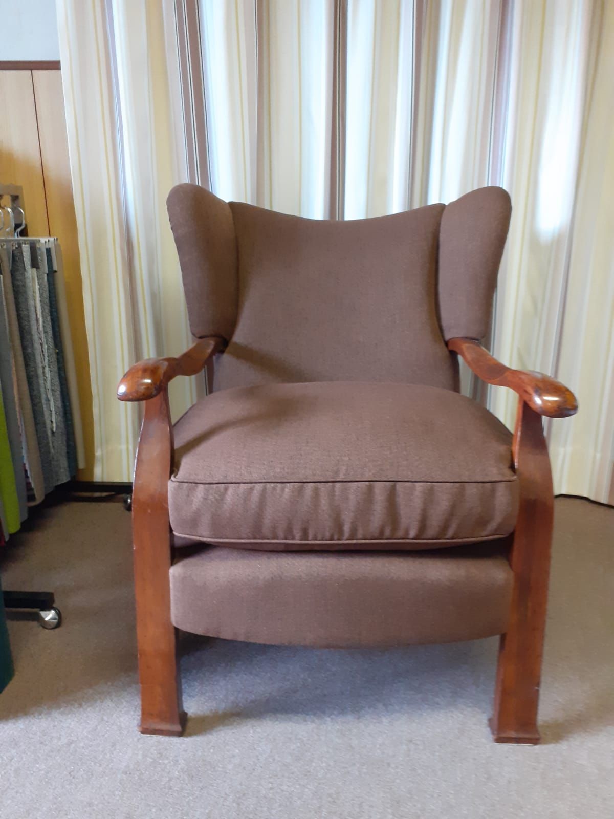 Poltrona con schienale alto imbottita marrone e struttura in legno, davanti a tende a righe.