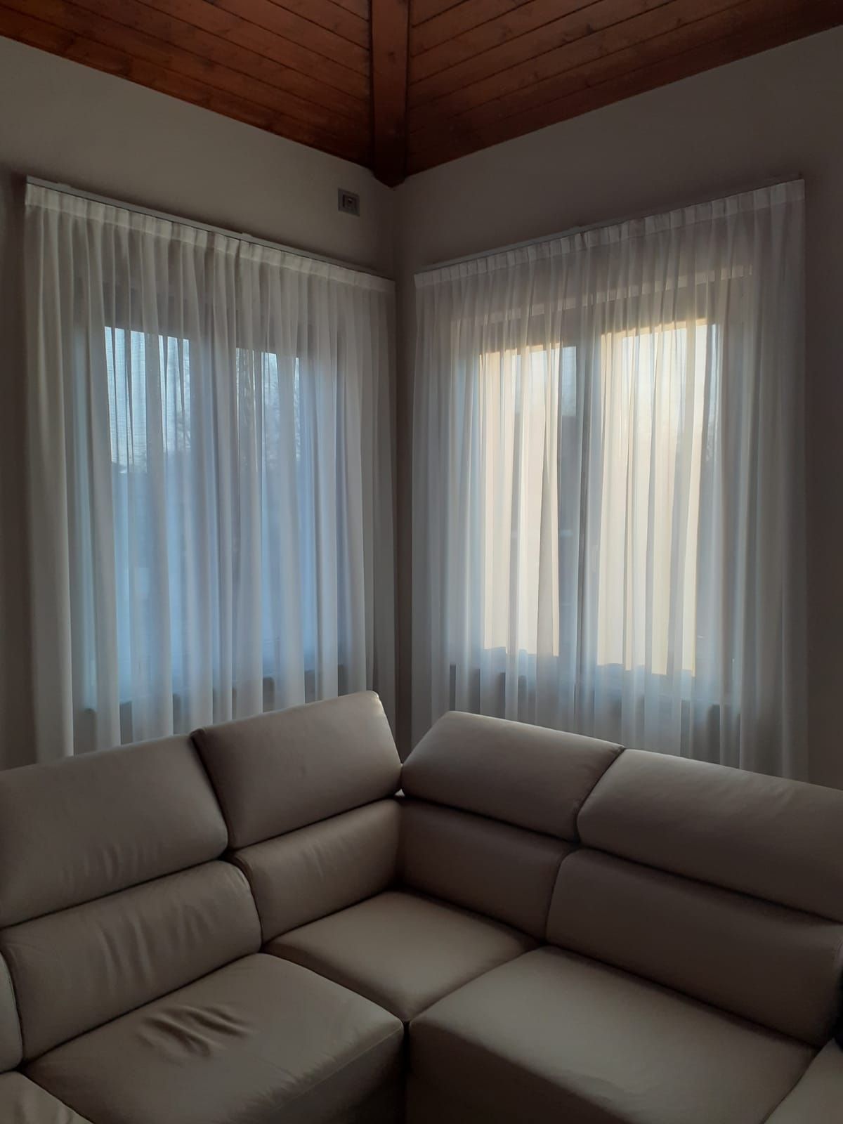 Angolo di una stanza con tende bianche trasparenti su due finestre sopra un divano componibile beige. La luce del sole entra a fiotti.