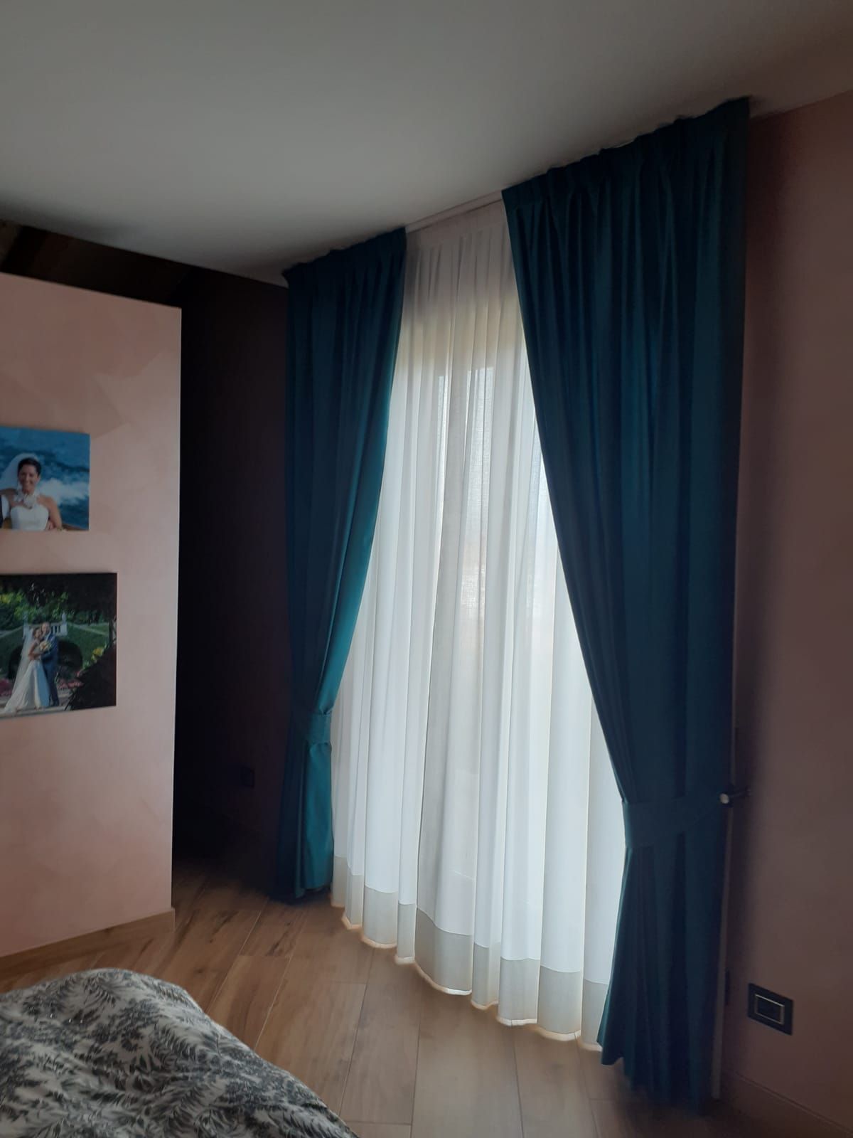 Tende color verde acqua incorniciano una finestra con tende bianche trasparenti. Una parete rosa con foto di nozze.