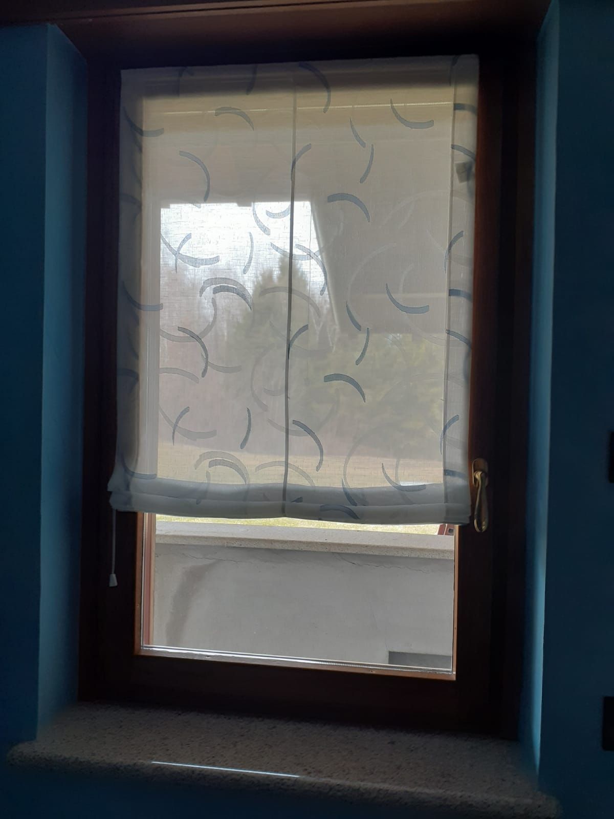 Finestra con una tenda trasparente e fantasia che copre parzialmente il vetro. Cornice marrone, parete blu.