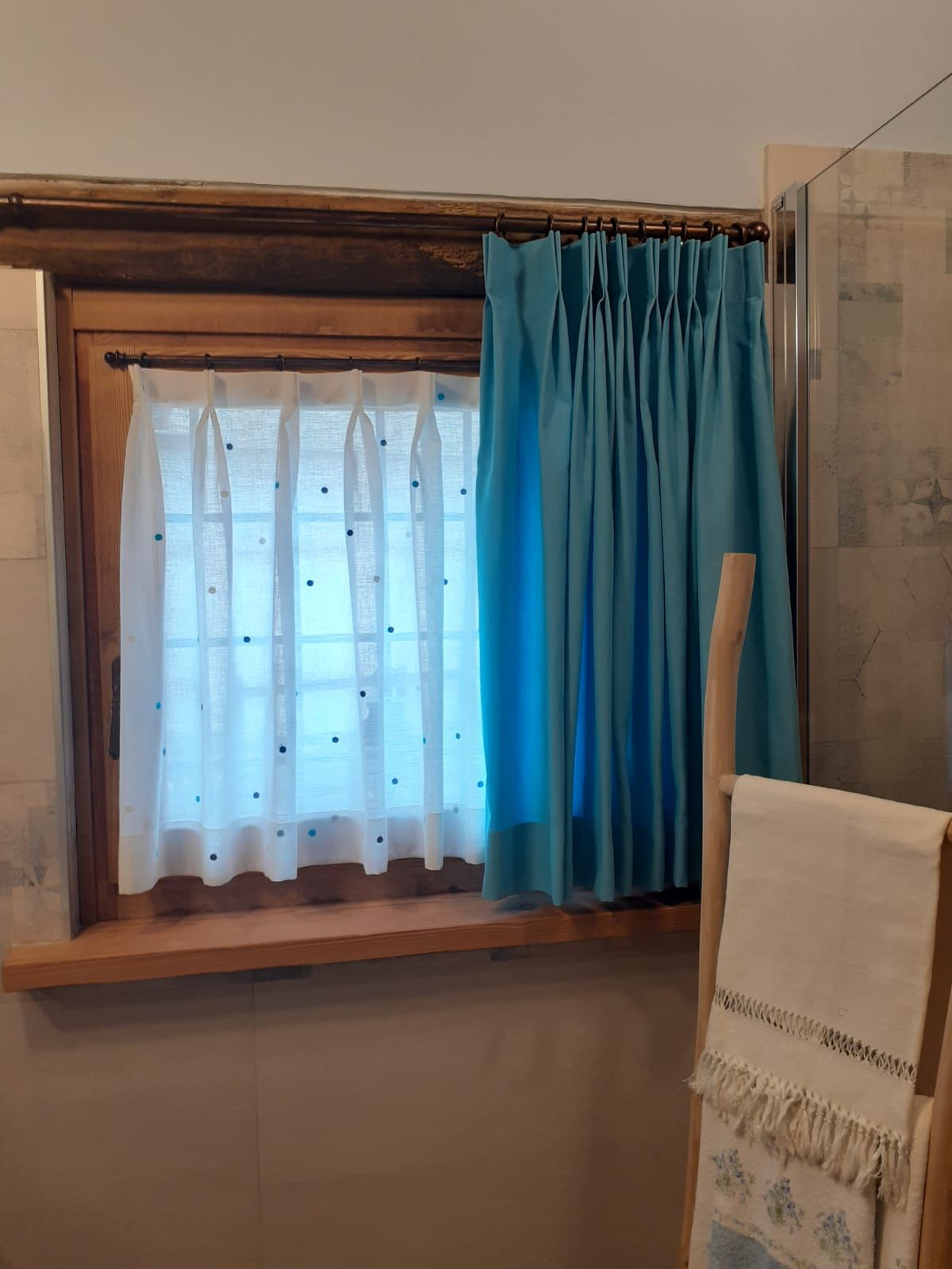 Finestra del bagno con tenda trasparente a pois, drappeggio blu e cornice in legno. Portasciugamani con asciugamano bianco.
