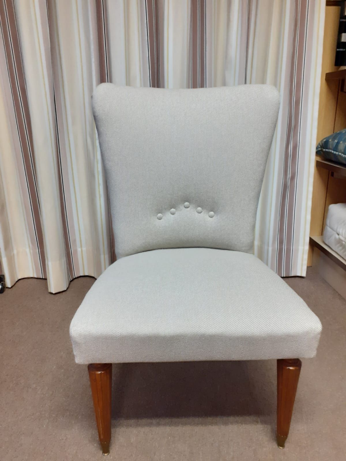 Sedia imbottita con tessuto grigio chiaro, gambe in legno e dettaglio con bottoni sullo schienale. Posizionata davanti a tende a righe.