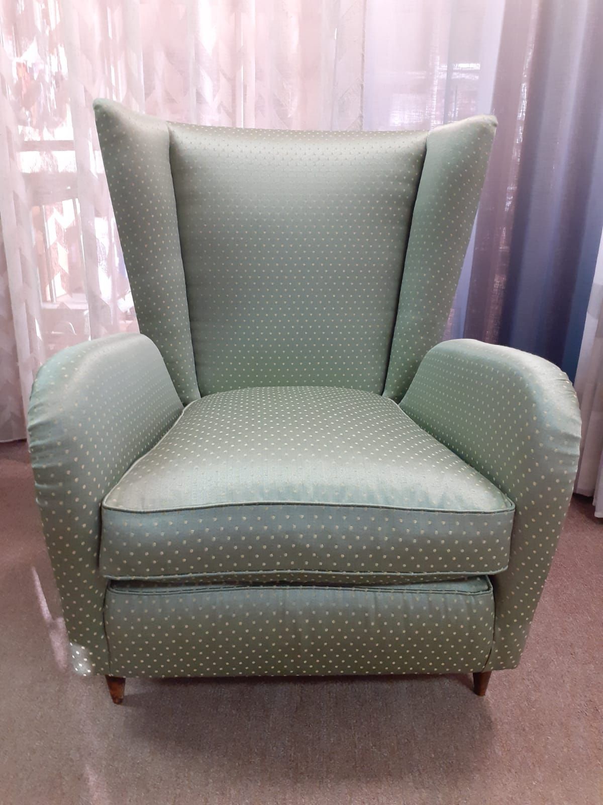 Poltrona verde chiaro con pois bianchi, schienale alto, gambe in legno, posta davanti a tende trasparenti.
