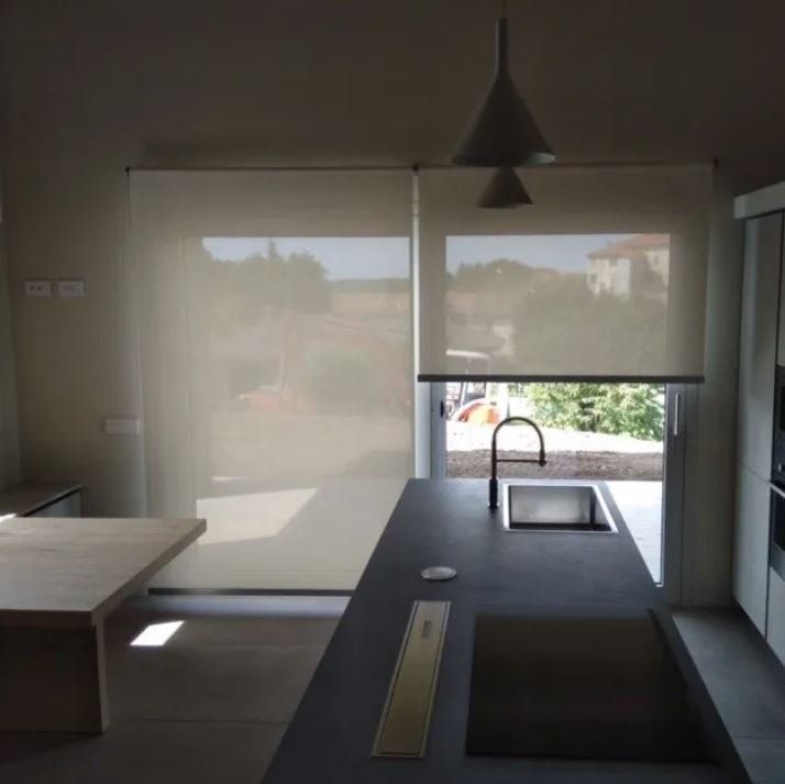 Cucina con tende a rullo di colore chiaro, piano di lavoro grigio scuro, lavello e vista sugli alberi.