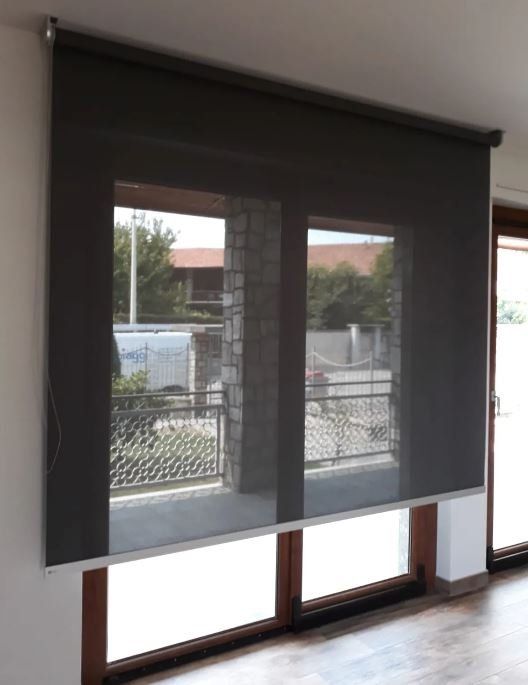 Una tenda avvolgibile grigio scuro copre due finestre, consentendo la vista su un balcone esterno.