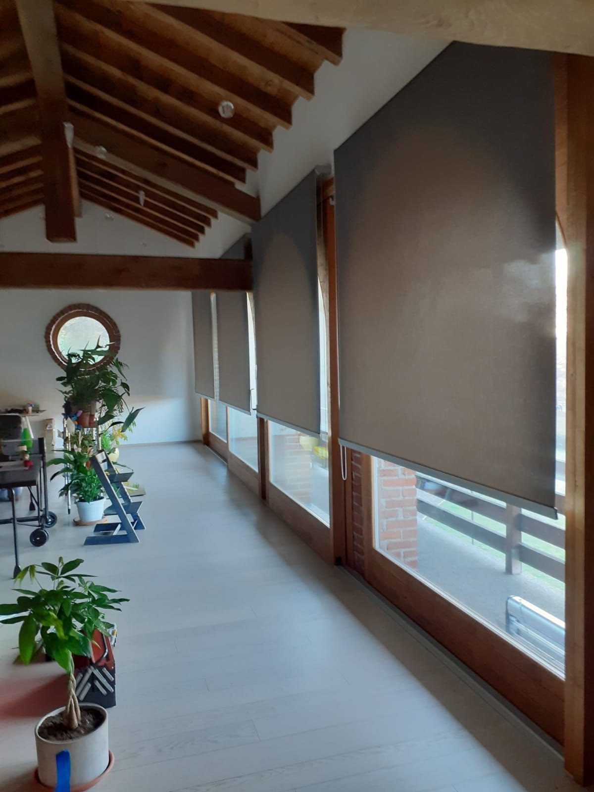 Vista interna di una stanza soleggiata con ampie finestre, tende a rullo grigio scuro, travi in ​​legno e piante da interno.