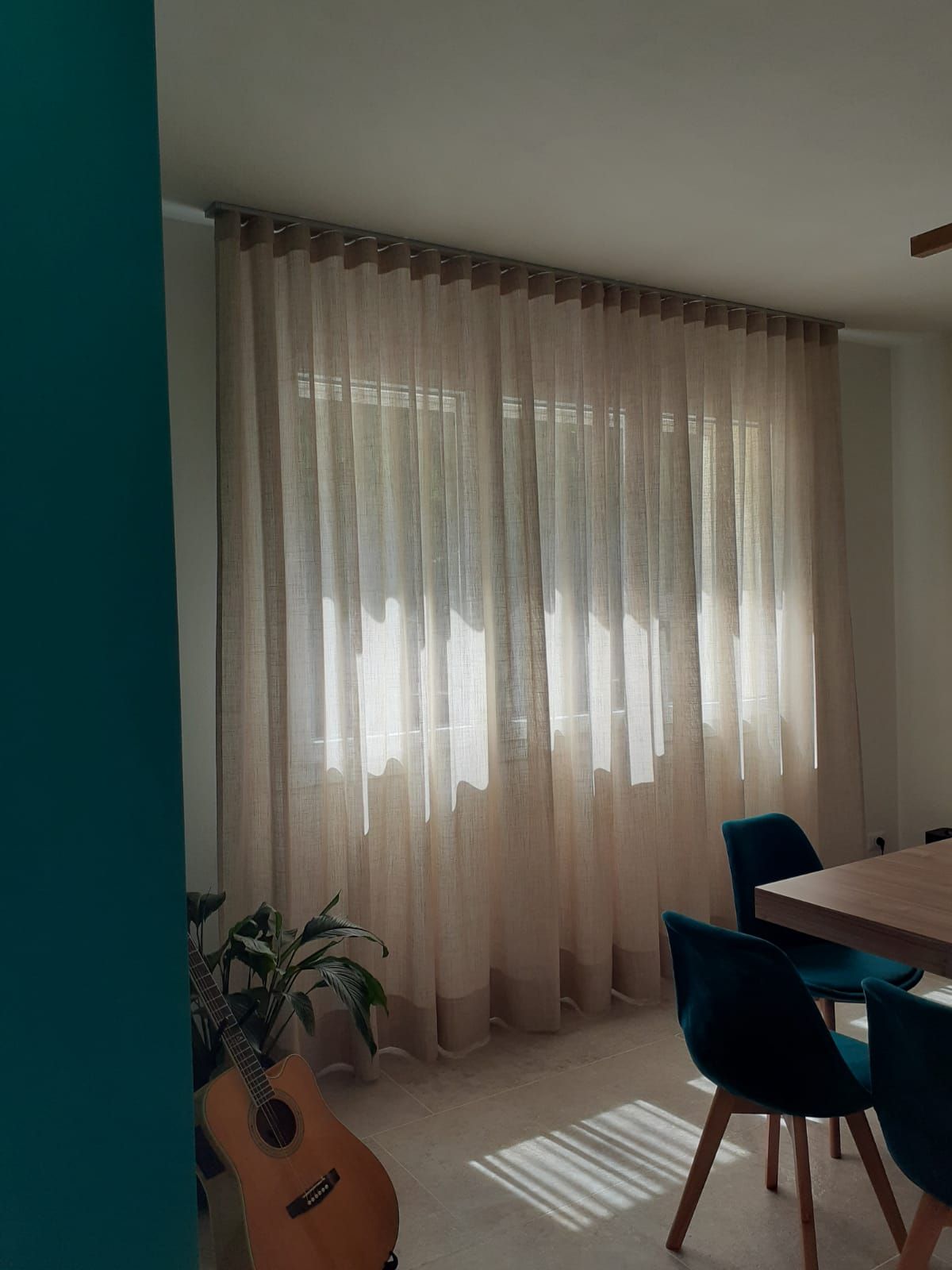 Tende beige trasparenti sopra una finestra in una sala da pranzo, che lasciano filtrare la luce del sole. Sedie color verde acqua su un tavolo di legno.