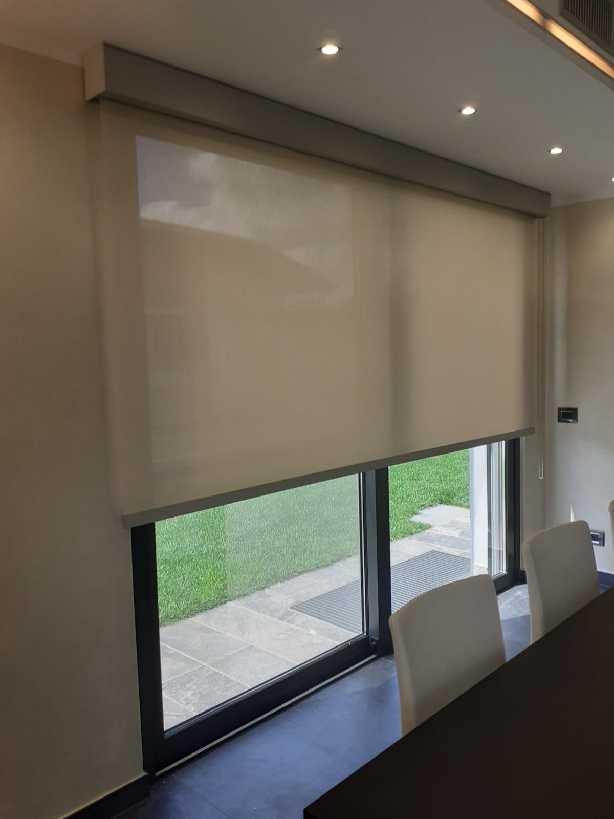 Tende a rullo beige che coprono una finestra con vista sul verde. All'interno di una stanza con tavolo e sedie.