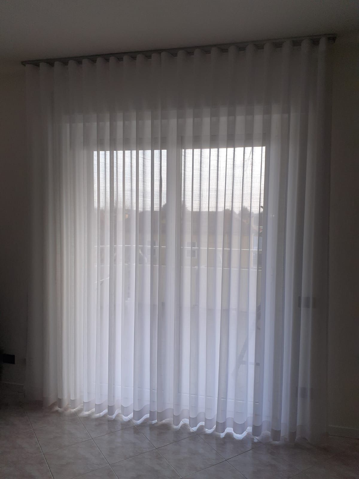 Tende bianche trasparenti appese davanti a una finestra, che proiettano ombre verticali.