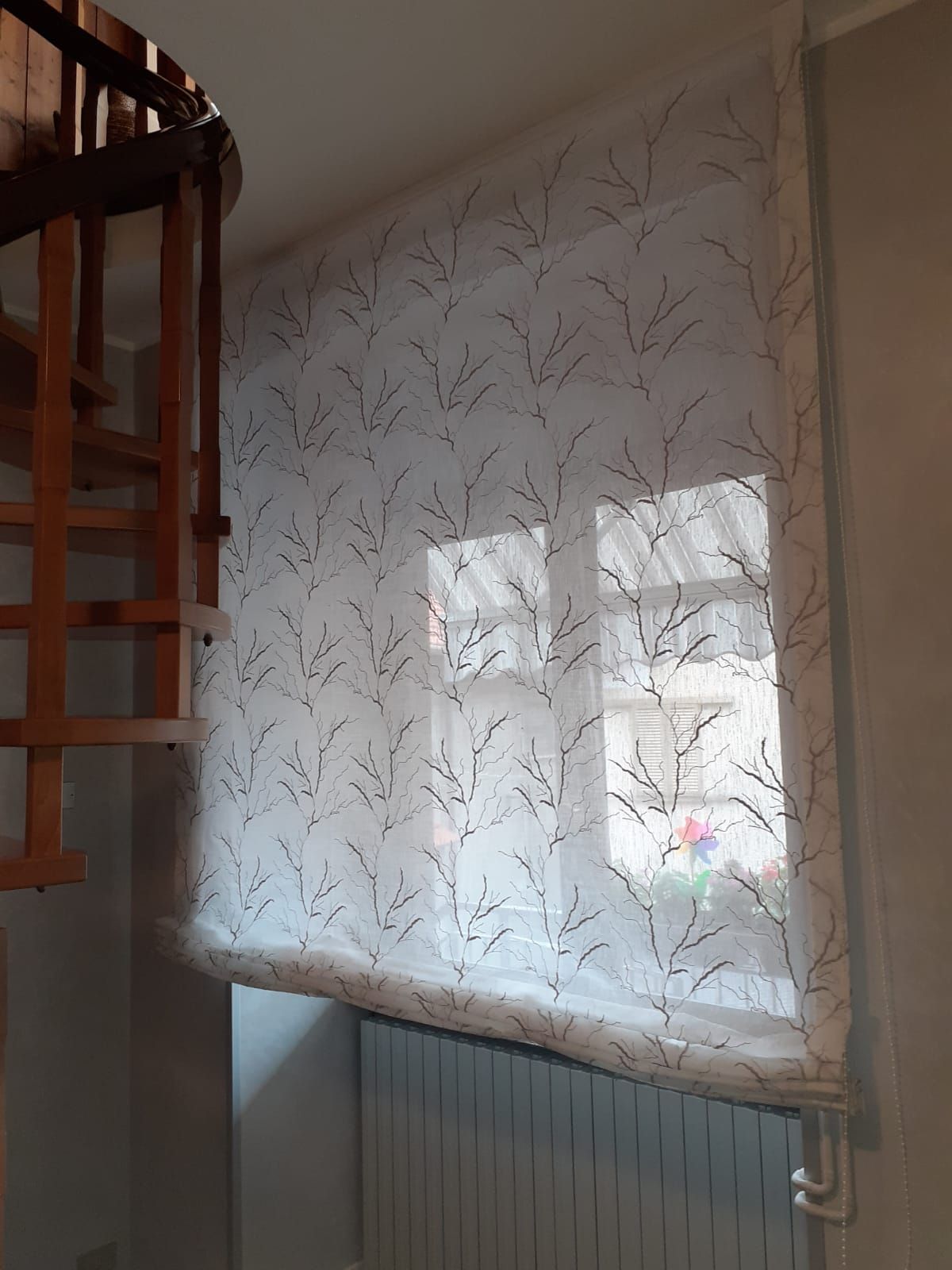 Tenda bianca con motivo a rami neri, che copre una finestra sopra un termosifone. È visibile una scala a chiocciola in legno.
