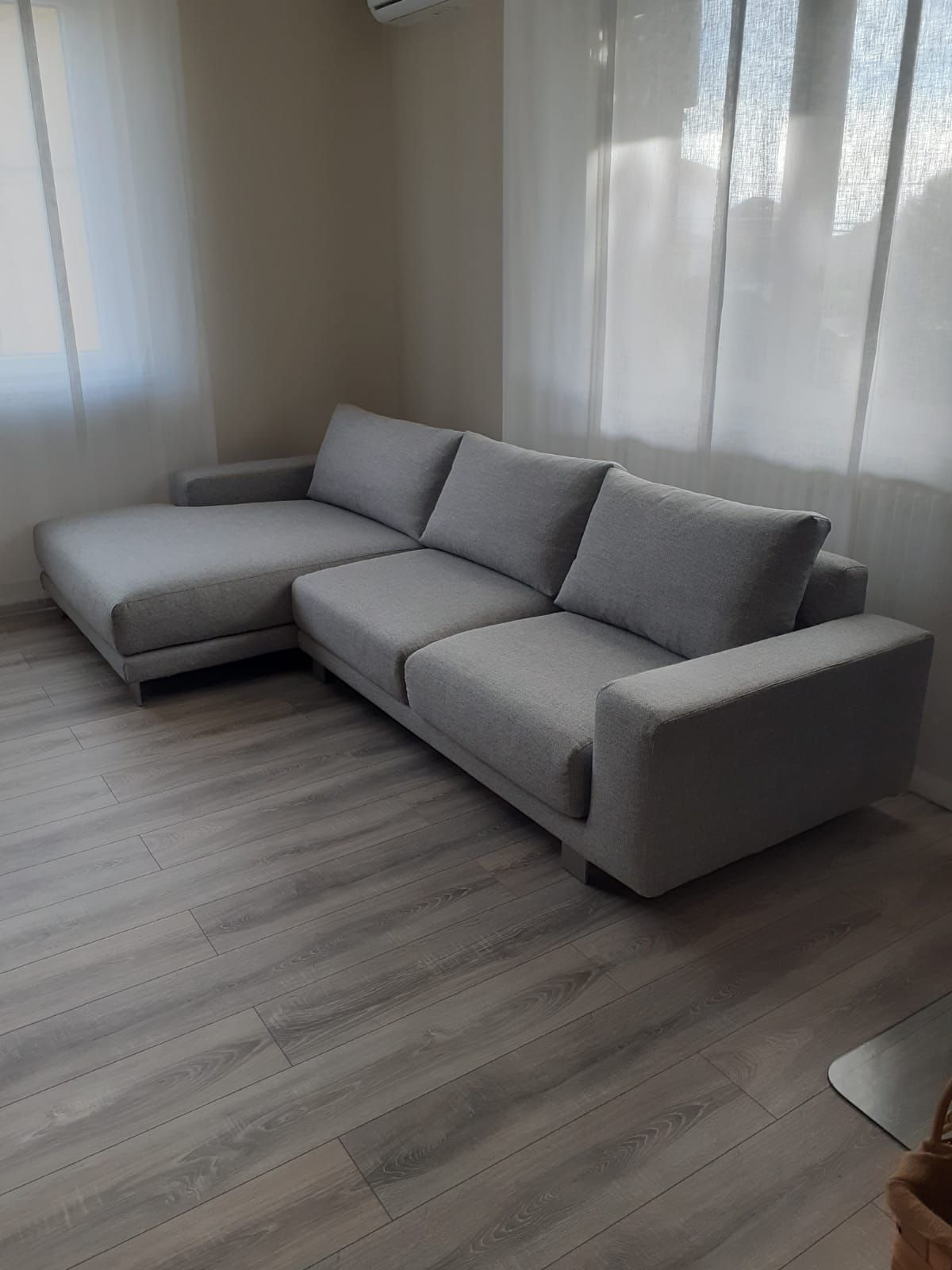 Divano componibile grigio in un soggiorno luminoso con pavimenti grigio chiaro e tende trasparenti.