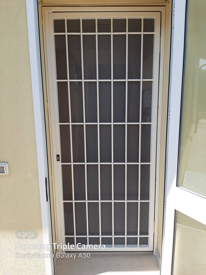 Cancello di sicurezza in metallo bianco sopra un ingresso color sabbia con pareti beige e un piccolo campanello sulla sinistra.