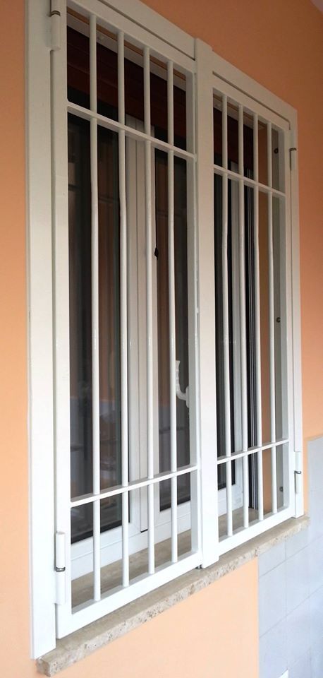 Finestra con sbarre bianche e grate di sicurezza su una parete esterna arancione.