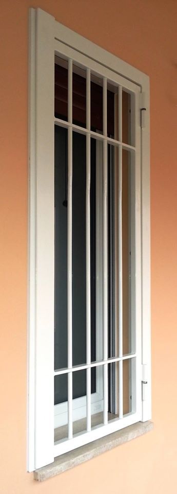 Finestra con sbarre bianche e cornici beige, dietro la quale si intravede un interno scuro parzialmente aperto.