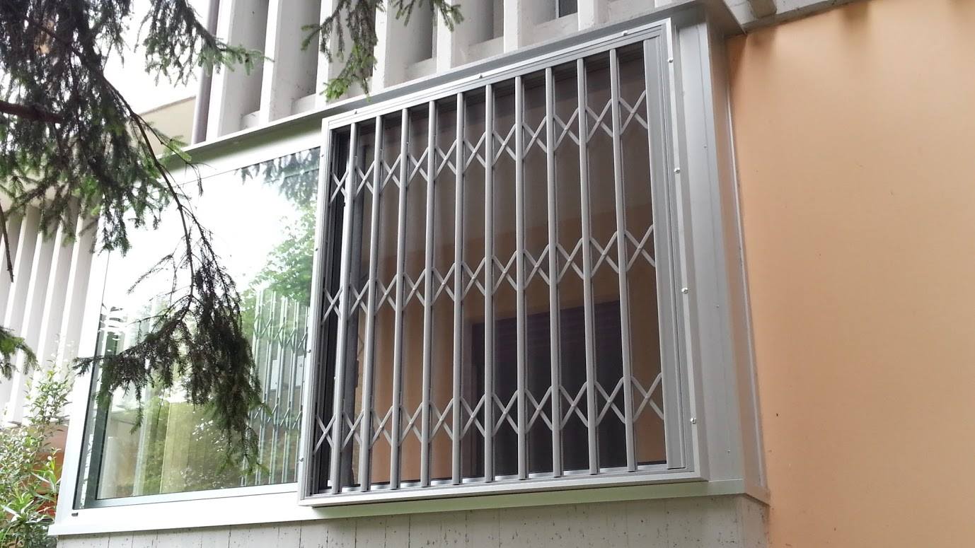 Finestra con sbarre bianche su un edificio beige con persiana marrone e vegetazione riflessa nel vetro.