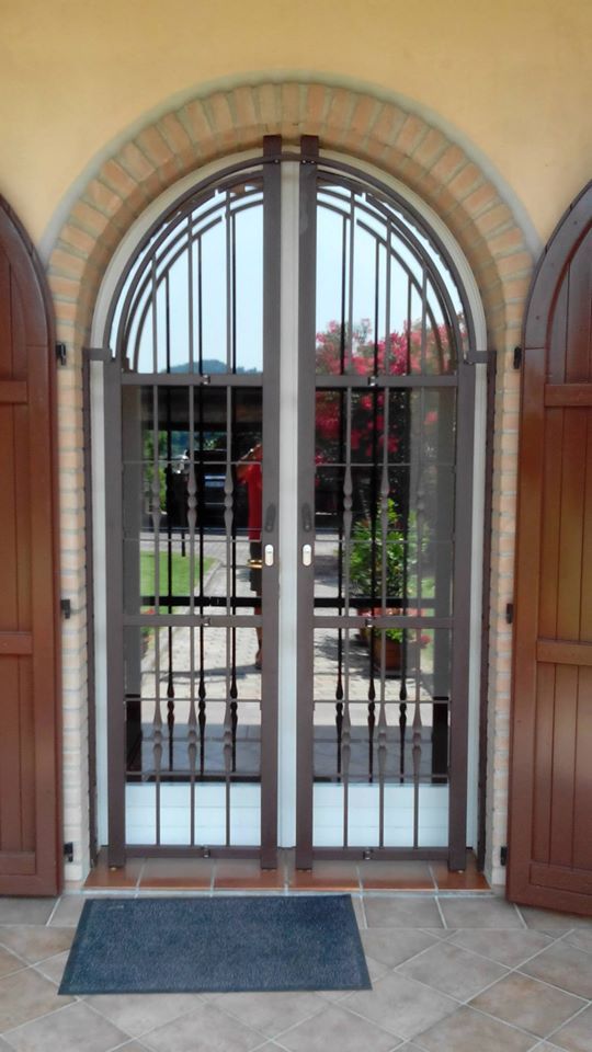Porta d'ingresso ad arco con porte in legno a vista e cancello di sicurezza in ferro battuto, che conduce all'esterno su un patio.