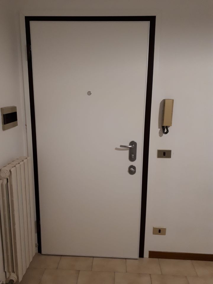 Porta d'appartamento bianca con finiture nere, maniglia argentata, citofono e termosifone in un corridoio