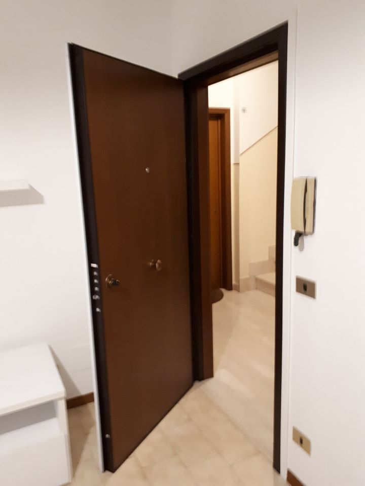 Apri la porta marrone dell'appartamento che dà su un corridoio illuminato, con un citofono a muro accanto.
