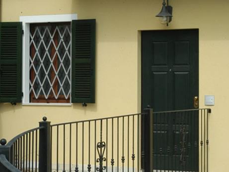 Facciata di casa gialla con porta verde scuro, finestra con persiane e ringhiera in ferro battuto nero.