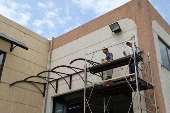 Operai su un'impalcatura installano una tettoia metallica sulla facciata di un edificio beige.