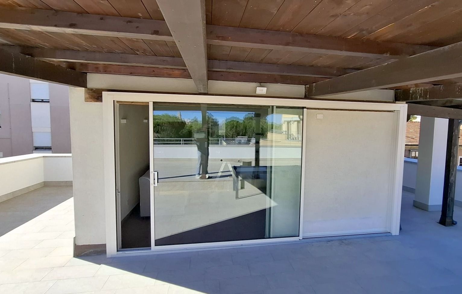 Patio coperto con porta scorrevole in vetro e pareti in stucco bianco, che si apre su un luminoso passaggio esterno.