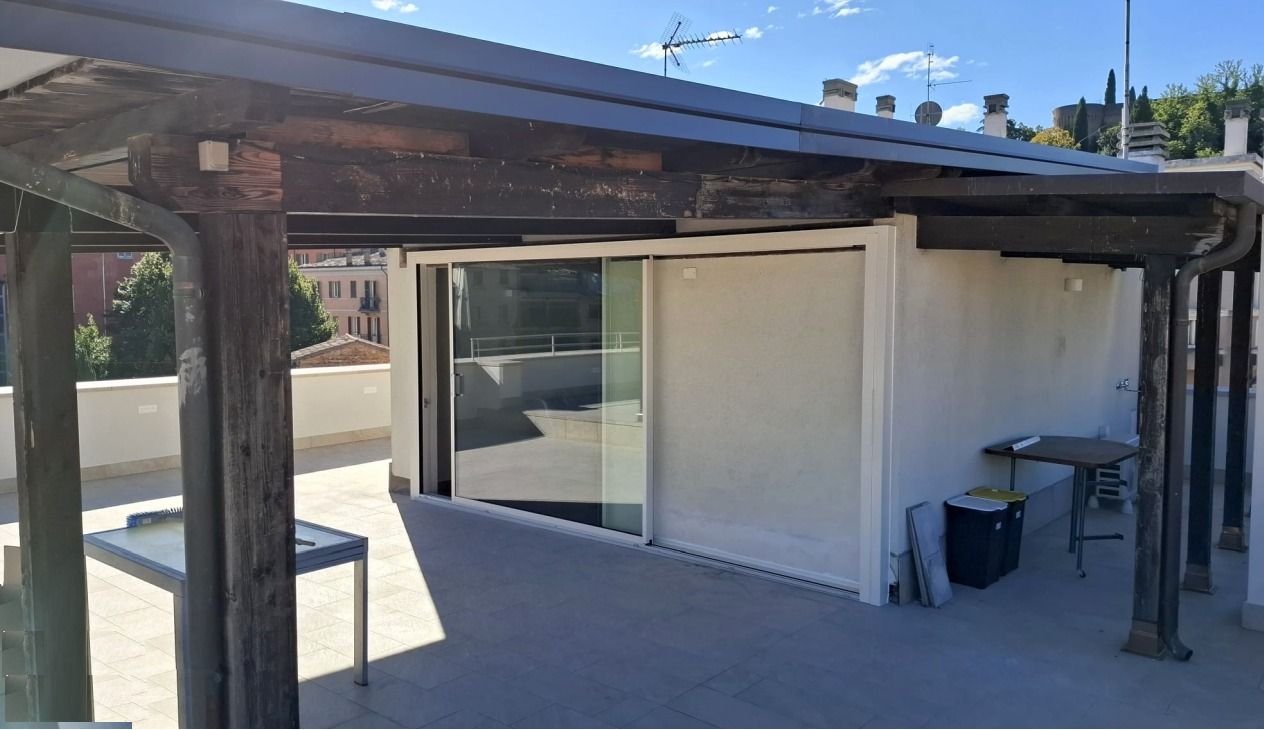 Patio con pergola in legno invecchiato, porta scorrevole in vetro e tavolino da esterno addossato a un muro in stucco.