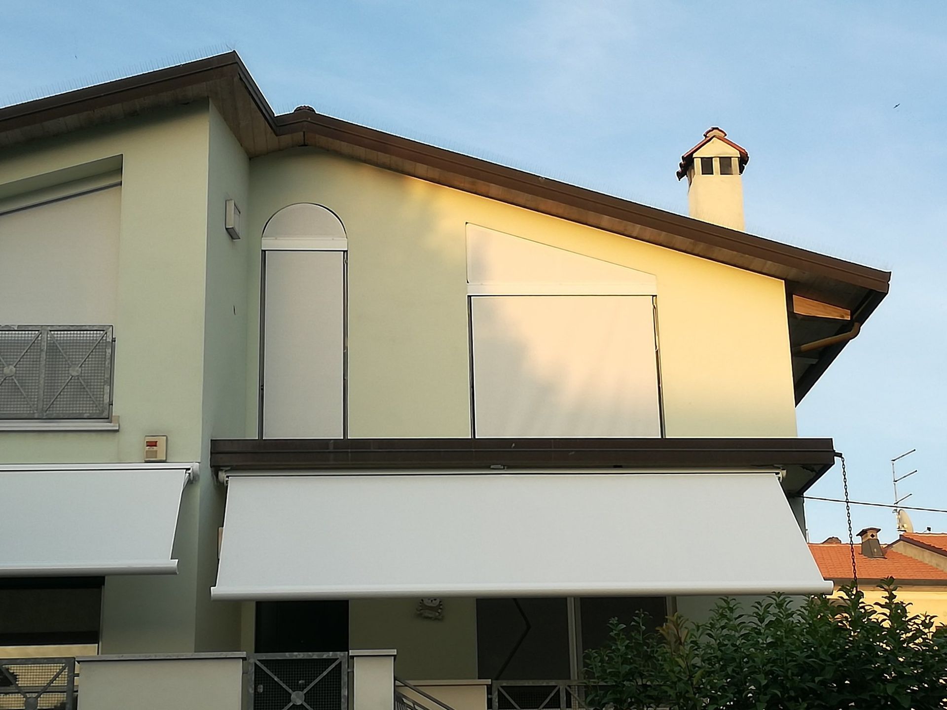 Esterno di una casa a due piani con pareti beige, tende da sole bianche e un piccolo camino al tramonto.