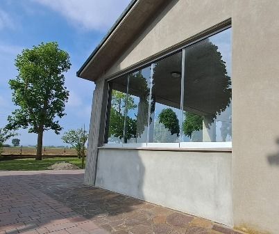 Esterno di un edificio moderno con ampie finestre, vialetto pavimentato e un albero accanto a un paesaggio aperto.