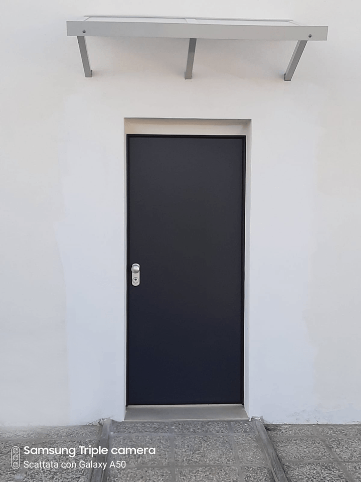 Muro di un edificio bianco con una porta scura e una piccola tettoia metallica sopra di essa.