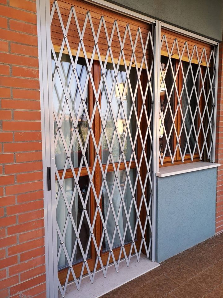 Cancello di sicurezza metallico chiuso sopra una porta a vetri e una finestra, accanto a un muro di mattoni.