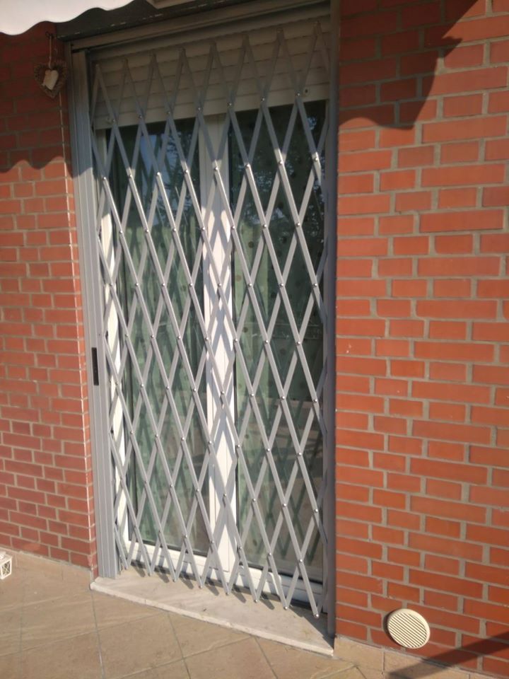 Esterno di una casa in mattoni con un cancello di sicurezza in metallo bianco sopra una porta a vetri, illuminato dal sole.