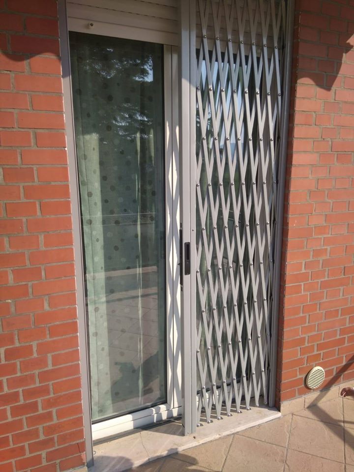 Esterno in mattoni con porta a vetri e cancello di sicurezza a soffietto color argento su un patio soleggiato.