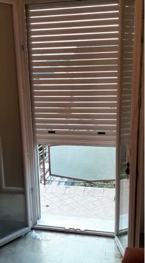 Tende bianche parzialmente aperte sopra una porta, con luce naturale e un piccolo patio esterno visibili dall'esterno