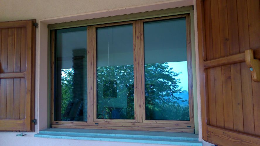 Finestra in legno con persiane aperte che riflette alberi e cielo sulla facciata di un edificio.